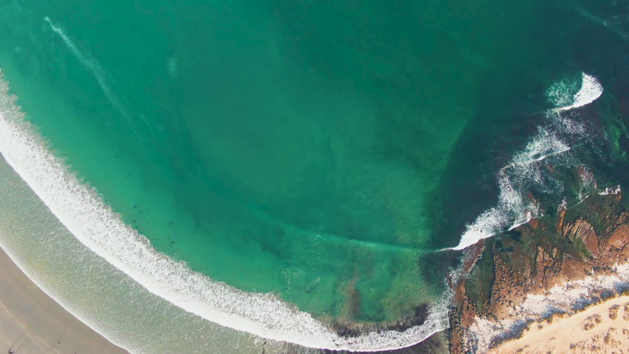 High overhead view in La Bocana with a bright foam band across turquoise water, exposed rocks on the right edge, sandy shore below, and no visible people.