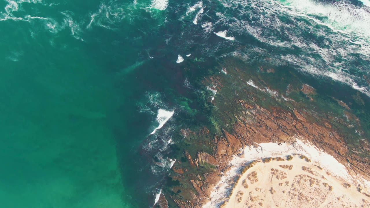 Aerial frame from La Bocana, Baja California Sur showing turquoise water on the left, white surf crossing dark rock, pale sand at the lower right, and no visible people.