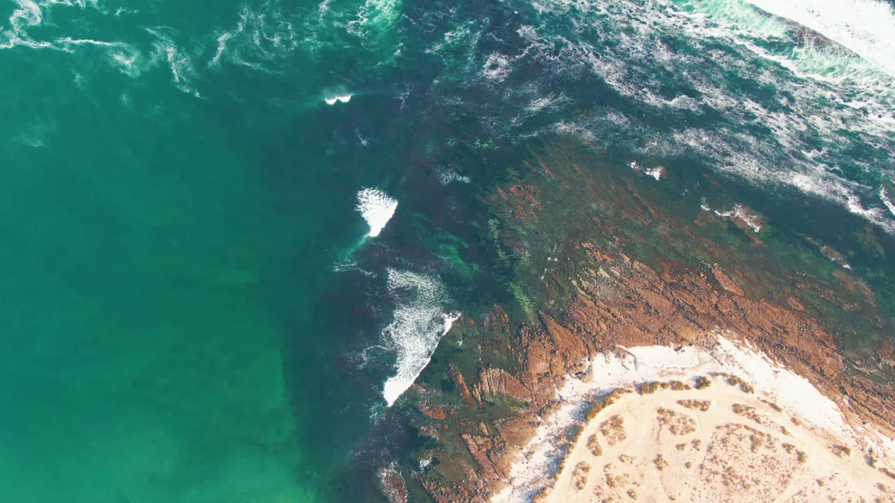 Aerial frame over La Bocana showing turquoise sea, white foam breaking on dark coastal rock, pale sand at the lower edge, and no visible people.