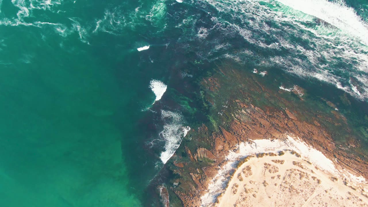 Overhead coastal water with surf lines crossing dark rocks and sand.