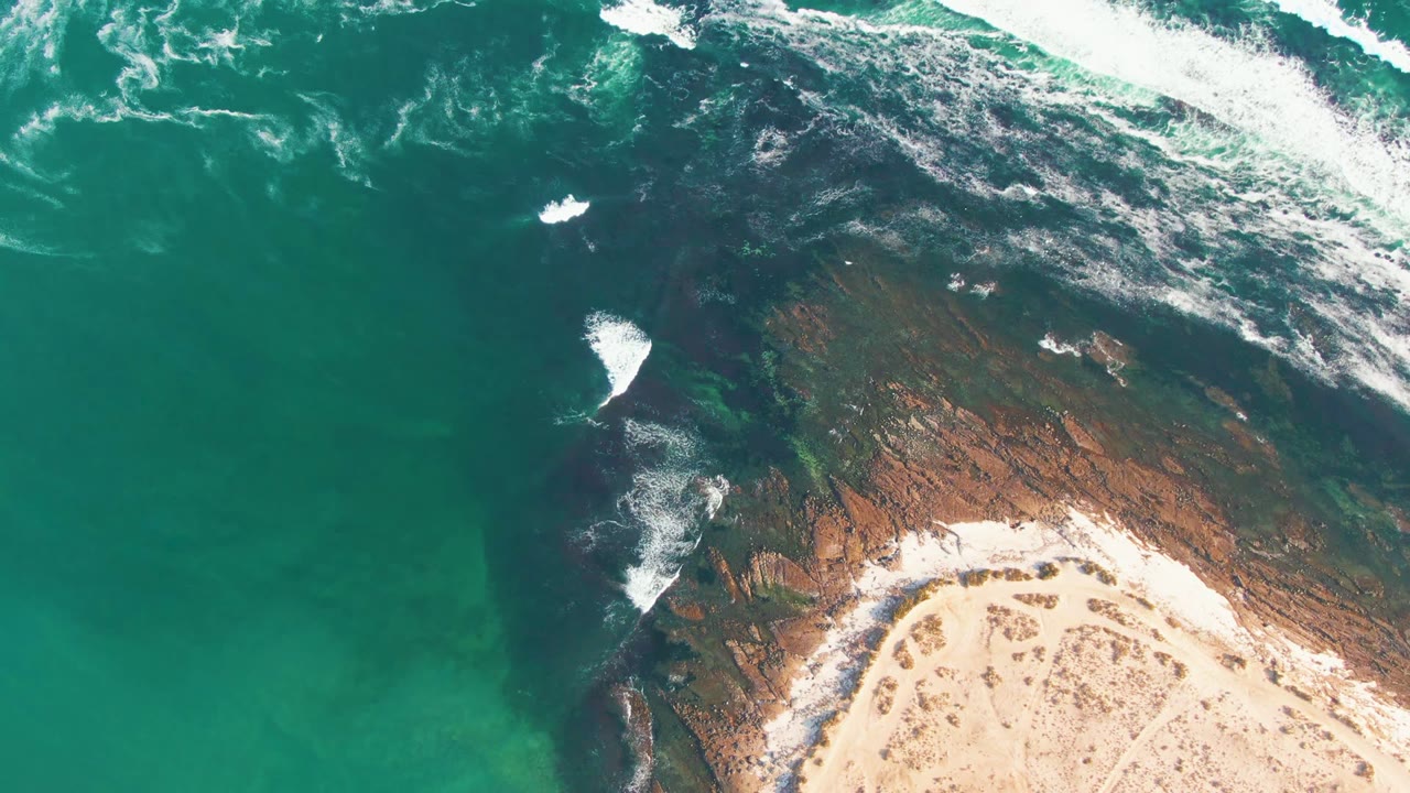Overhead frame of La Bocana coastline with white wave crests near the upper edge, foam on dark rock, curved sand at lower right, and no visible people.