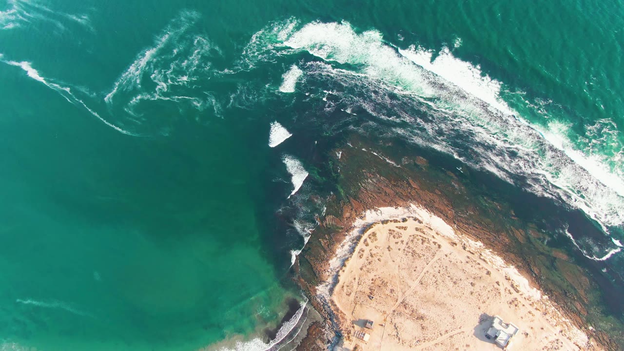 Overhead view of La Bocana, Baja California Sur coastline with turquoise water, white foam on dark rocks, sandy beach at the lower edge, and no visible people.