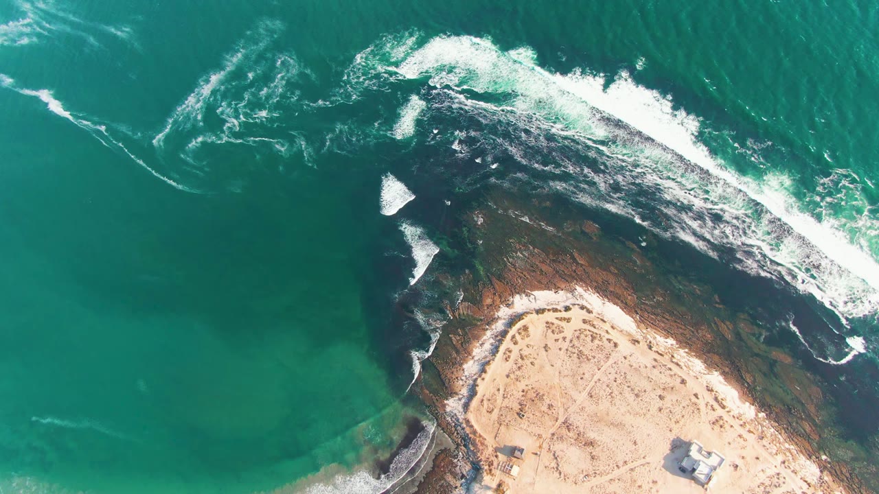 Overhead La Bocana coastline view with the foam line shifted along the upper right rocks, teal water to the left, pale sand at the bottom, and no visible people.