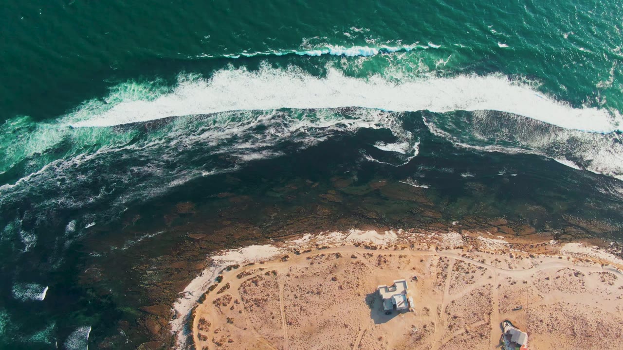 Aerial frame over La Bocana, Baja California Sur, with teal ocean, a white surf line, dark shallow rock or reef, sandy beach, small structures near the lower edge, and no visible people.