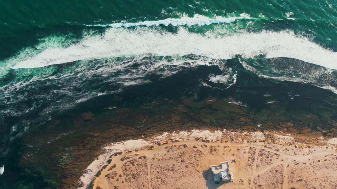Aerial La Bocana frame with a continuous white wave crest near the top, mottled dark shallows through the center, tan sand below, one prominent beach structure, and no visible people.