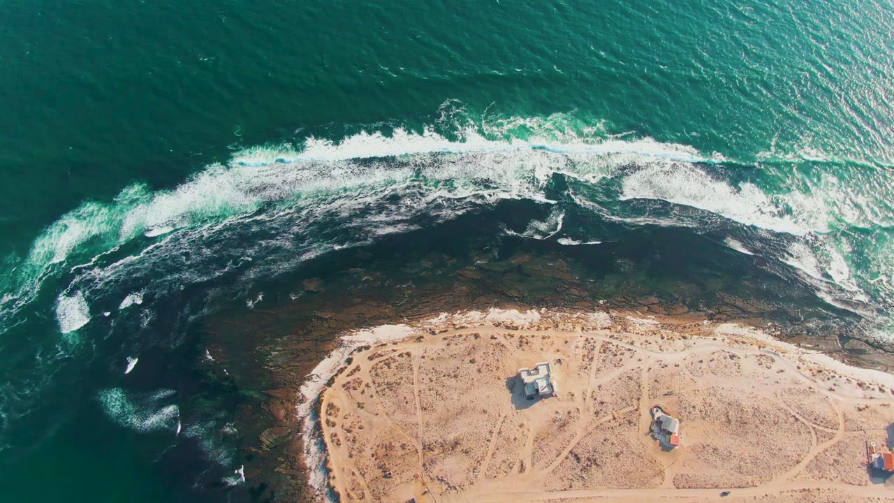 Opening overhead frame of La Bocana, Baja California Sur, with turquoise water, white surf over dark rocks, sandy shore, small structures, a red vehicle near the edge, and no visible people.