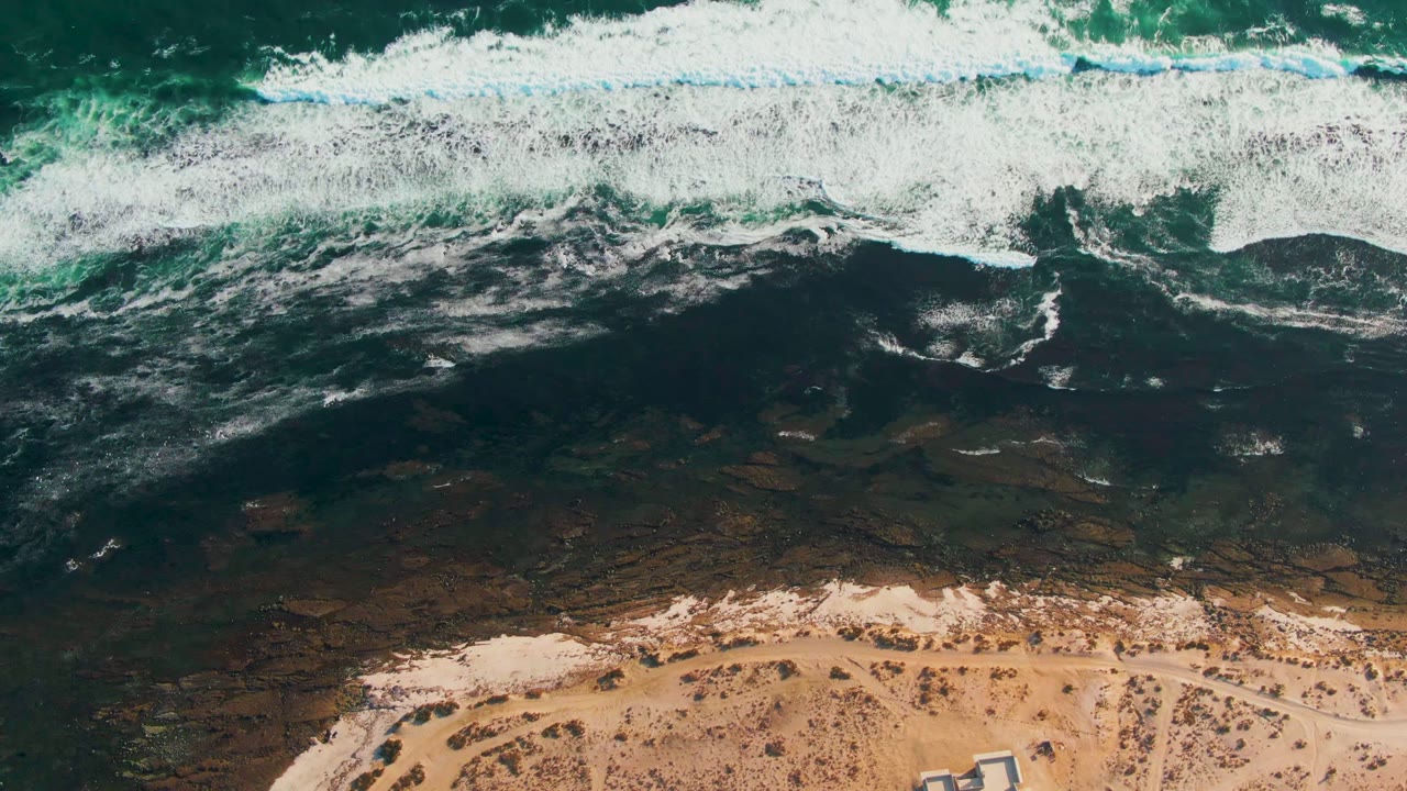 Overhead view of white surf breaking over dark coastal water toward a sandy La Bocana beach, with sparse vegetation near the bottom and no visible people.