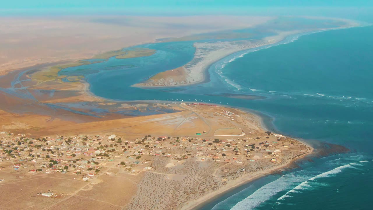 Opening aerial frame over La Bocana, Baja California Sur, with a small desert village in the lower frame, turquoise lagoon and pale sandbar beyond it, ocean at right, and no visible people.