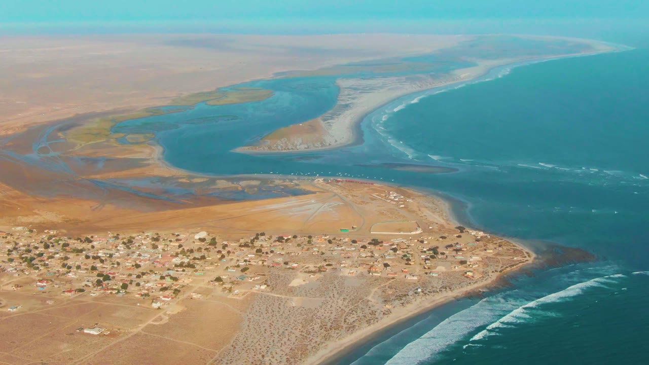 Desert settlement below a lagoon, sandbars, surf, and blue ocean.