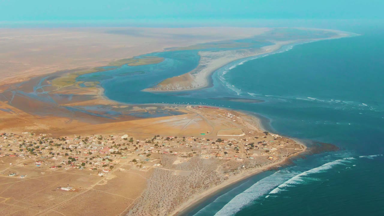 Wide aerial over La Bocana with the village spread across the lower left, a pale sand spit between lagoon and ocean, white surf lines along the coast, and no visible people.