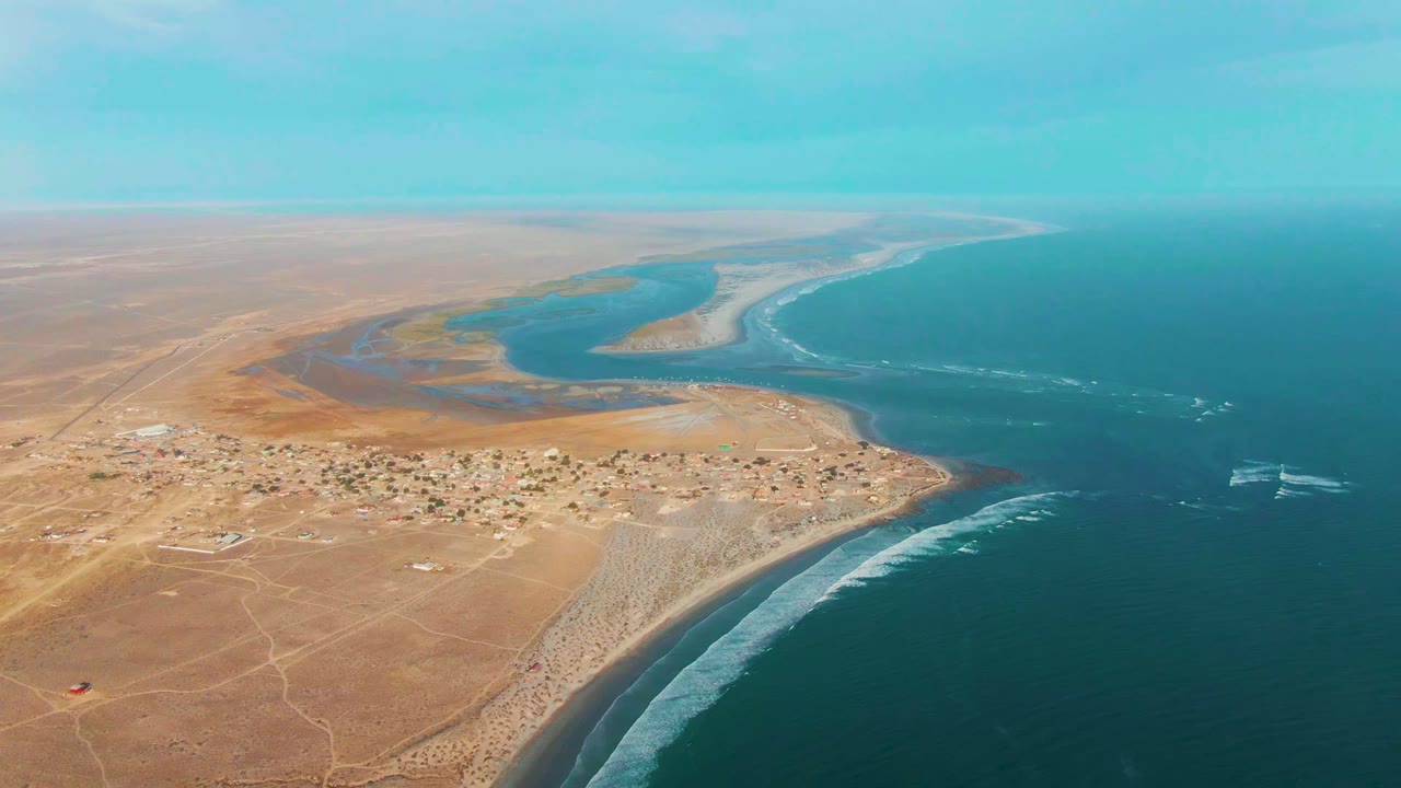 Aerial frame over La Bocana, Baja California Sur showing tan desert, a small shore settlement, river estuary, blue ocean, surf, and a small boat wake offshore, with no visible people.