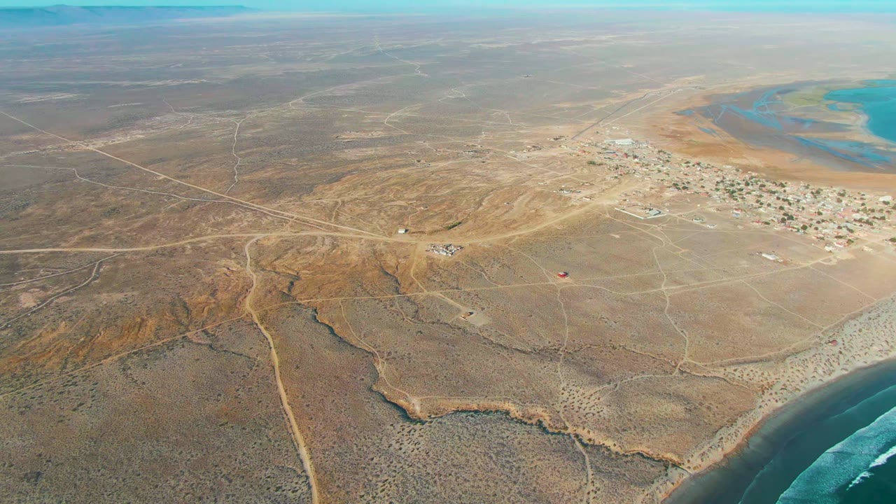 High aerial view over La Bocana, Baja California Sur, showing dry desert terrain, a small settlement near blue coastal water, pale dirt roads, and no visible people.