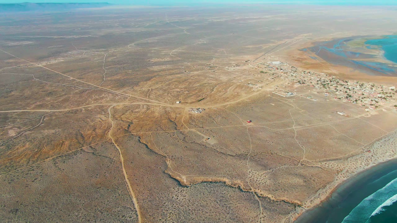 La Bocana aerial view with sandy ground in the foreground, a compact settlement in the distance, blue coastal water to the upper right, surf at the lower right, and no visible people.