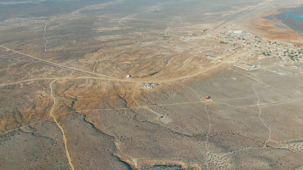 Aerial frame over La Bocana, Baja California Sur showing dry beige terrain, winding dirt tracks, scattered small buildings, a distant settlement, and no visible people.
