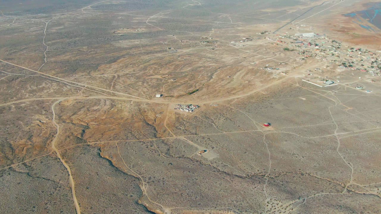 Wide drone view of arid terrain, branching dirt roads, and scattered structures near a settlement.