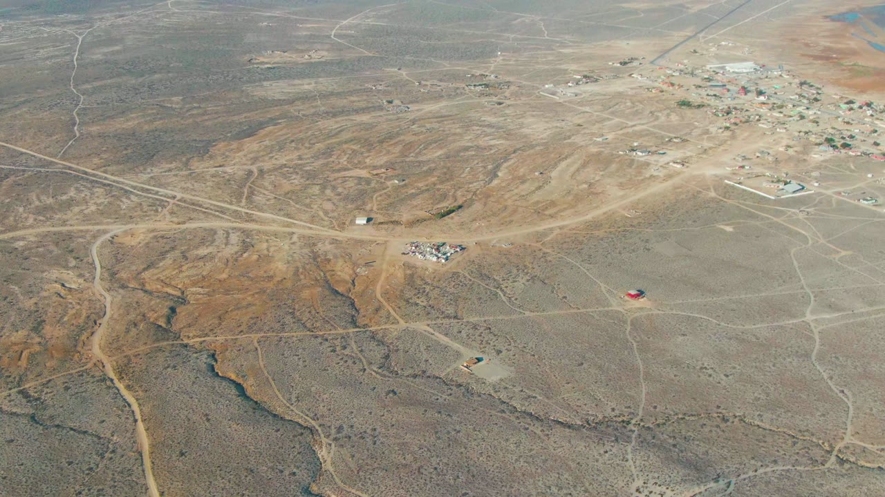 Drone-style aerial frame over La Bocana, Baja California Sur showing a compact settlement high in frame, pale dirt roads across dry terrain, a small red structure below center, and no visible people.