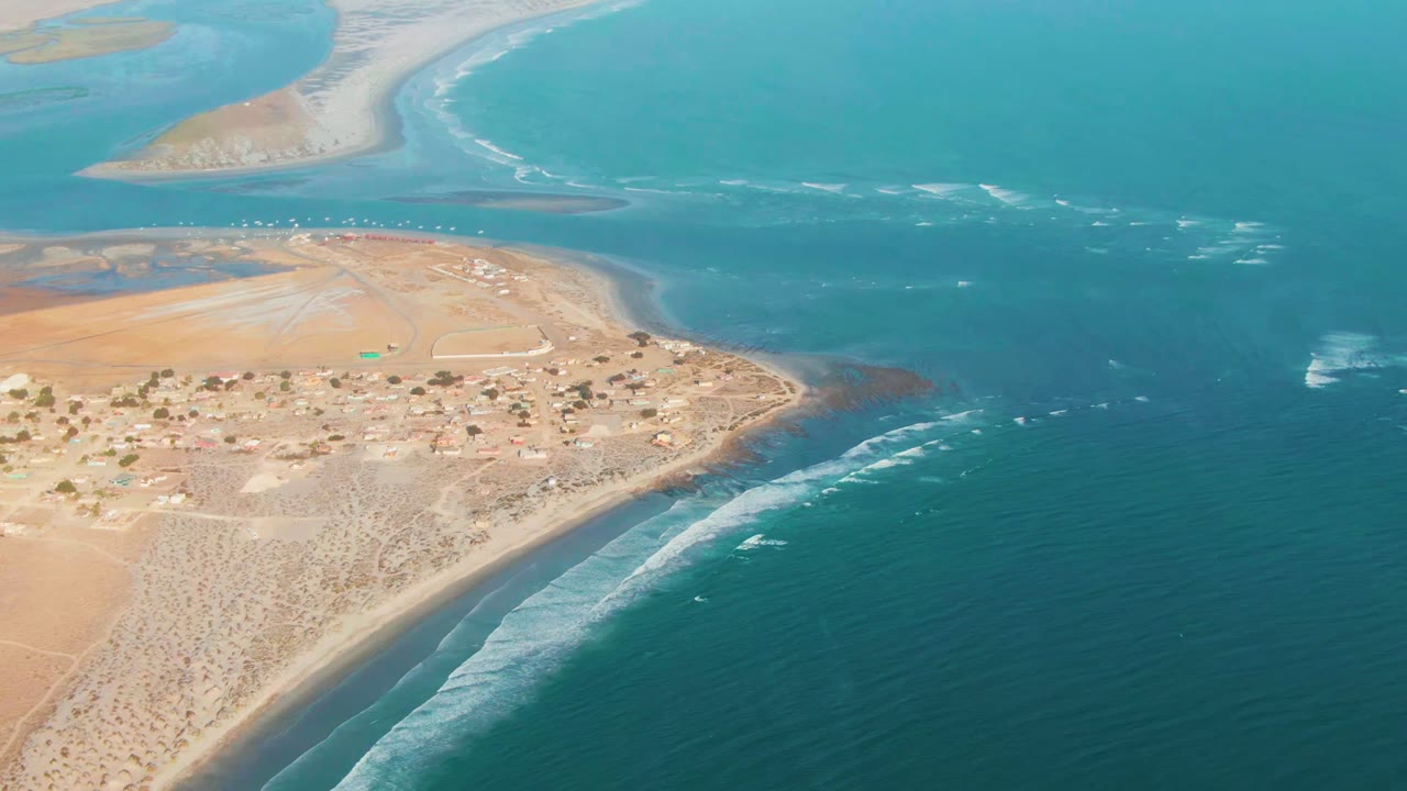 Aerial frame over La Bocana, Baja California Sur, showing sandy desert terrain, scattered structures, a curved shoreline, turquoise water, and no visible people.