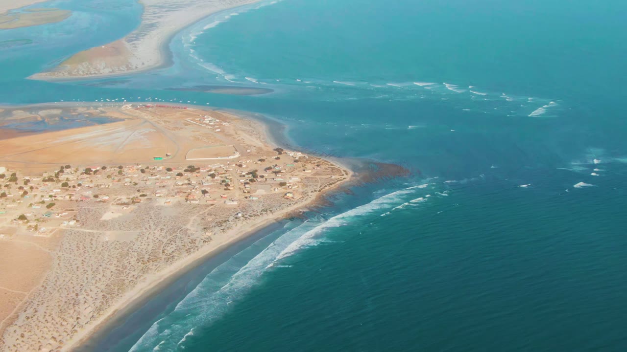 Aerial frame over La Bocana, Baja California Sur, showing tan desert land, a small shore settlement, blue ocean, an inlet, sandbars, and no visible people.