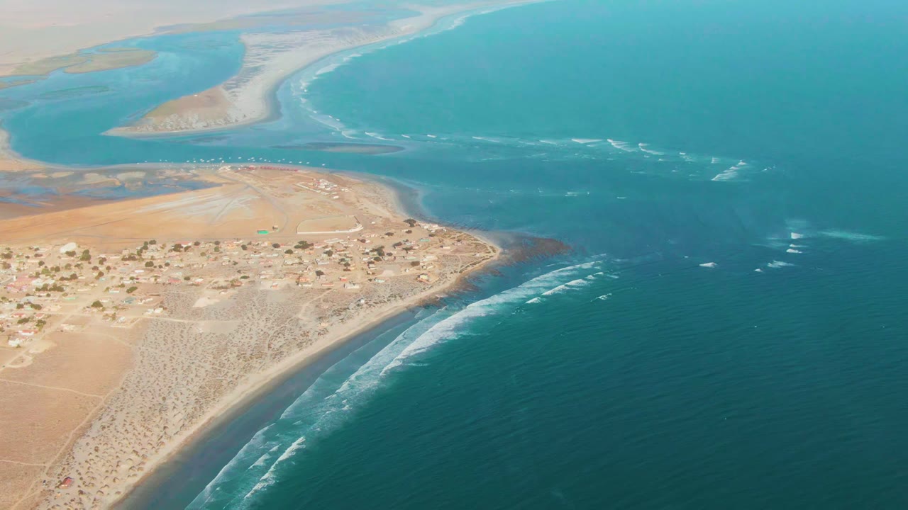 Drone-height view over La Bocana with arid ground and low buildings at left, white wave lines along the coast, open blue water at right, and no visible people.