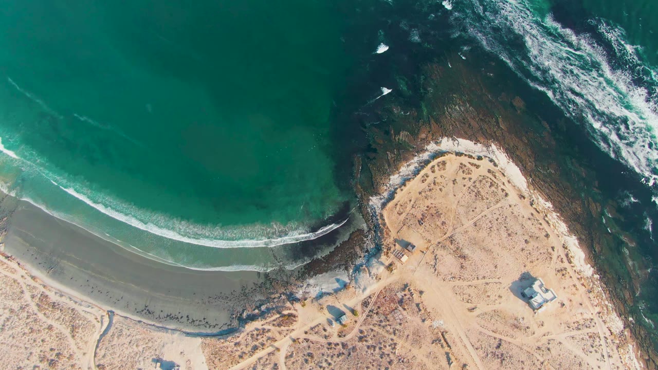 Aerial frame over La Bocana, Baja California Sur showing turquoise water, a curved line of white surf, sandy beach, dark rocks, small shore structures, and no visible people.