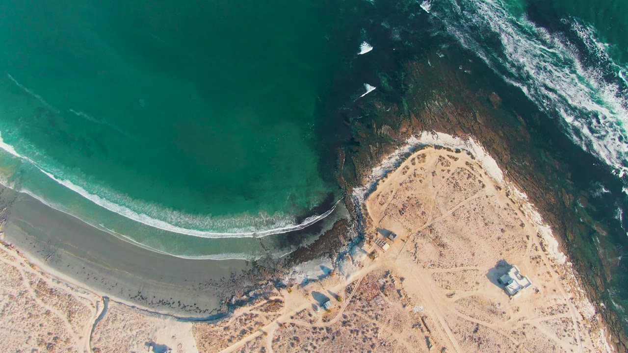 Top-down coastline with waves curling toward sand beside dark rocks.