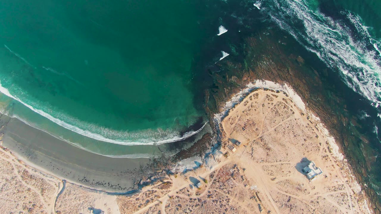 Aerial view of the La Bocana shore with green water at top, white foam crossing the wet sand, tan beach, small structures, and no visible people.