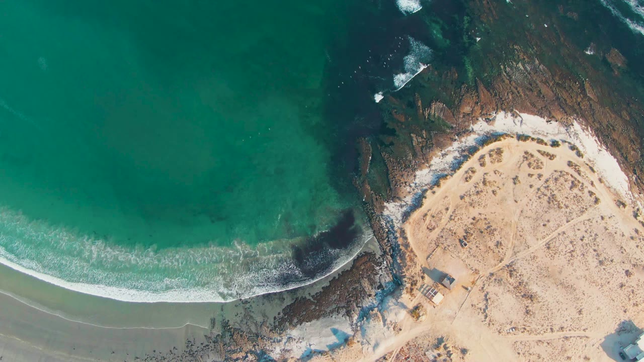 Overhead frame of La Bocana, Baja California Sur showing turquoise water on the left, white surf crossing the beach, dark rocks near center, pale sand on the right, and no visible people.