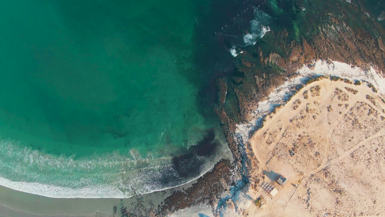 Top-down frame from La Bocana, Baja California Sur showing the beach shifted farther right in the composition, with turquoise shallows, broken foam, dark rocks, pale sand tracks, and no visible people.