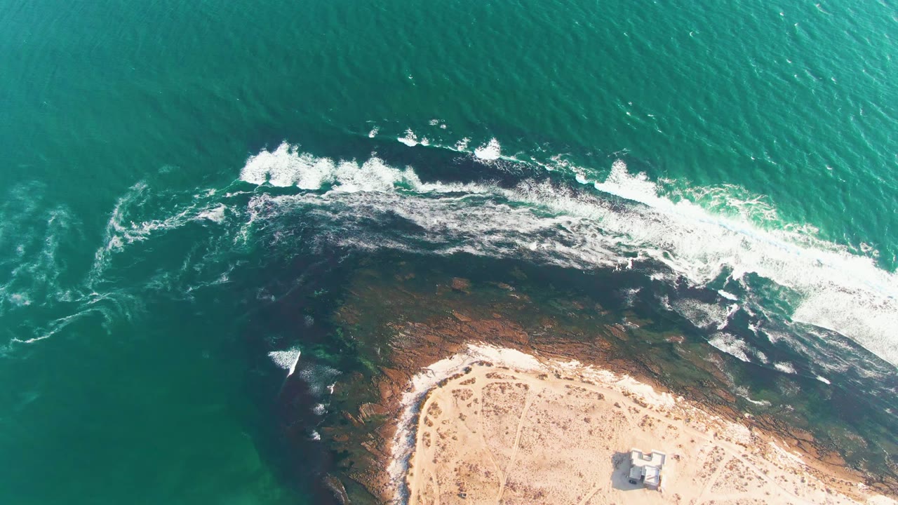 Overhead aerial frame of La Bocana, Baja California Sur, with turquoise ocean above, white foam crossing dark rock, pale sand below, a small structure near the lower edge, and no visible people.