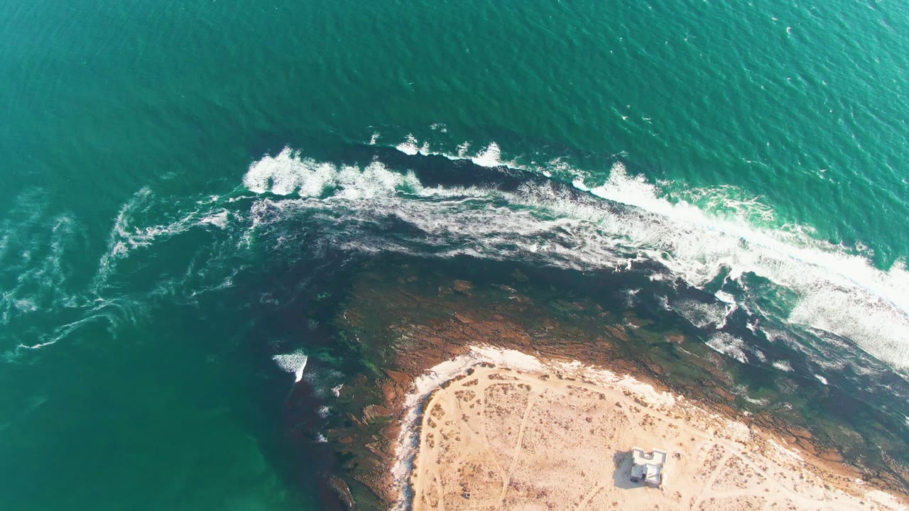 White surf spreads along a rocky reef with sand and a small structure below.
