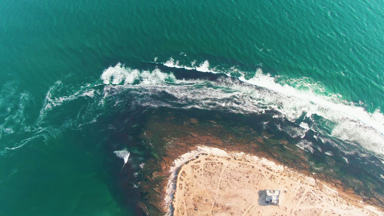 Top-down La Bocana frame where the breaking wave forms a bright white band over the rocky shoreline, with open turquoise water above and empty sandy beach below.
