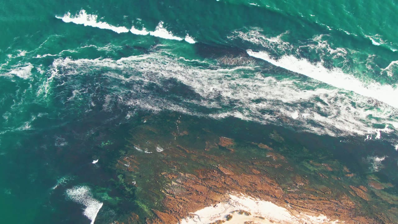 Aerial frame from La Bocana, Baja California Sur showing turquoise water, white surf, dark rock shelves, pale sand at the bottom edge, and no visible people.