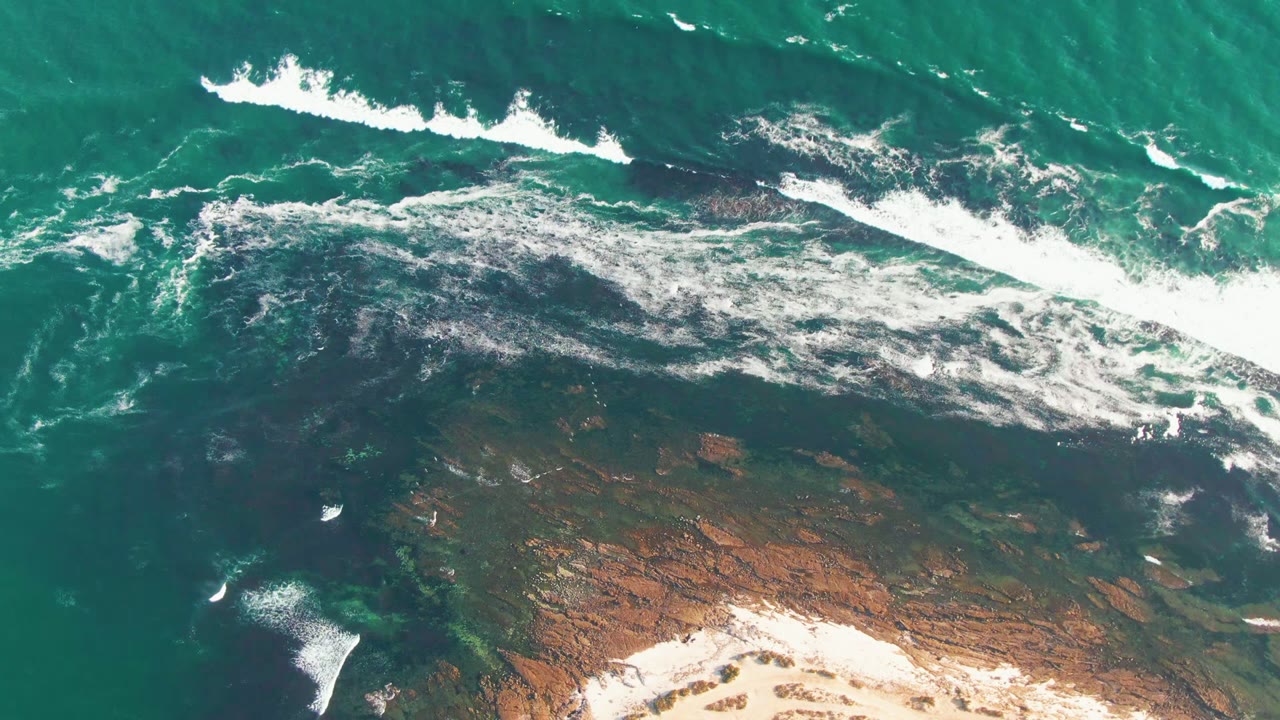 White wave foam stretches diagonally across shallow turquoise water and rocks.