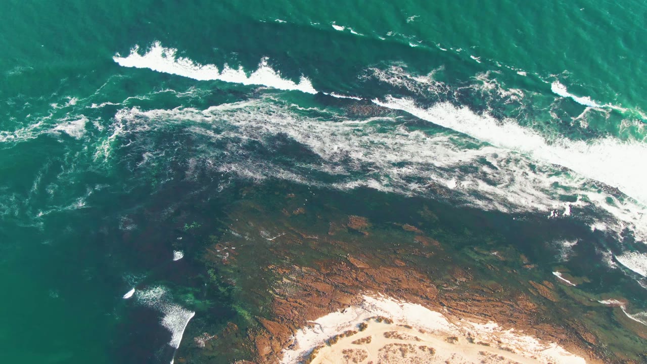 At 0:02, whitewater spreads farther across the dark rock shelf at La Bocana, with turquoise ocean above, pale sand below, and no visible people.