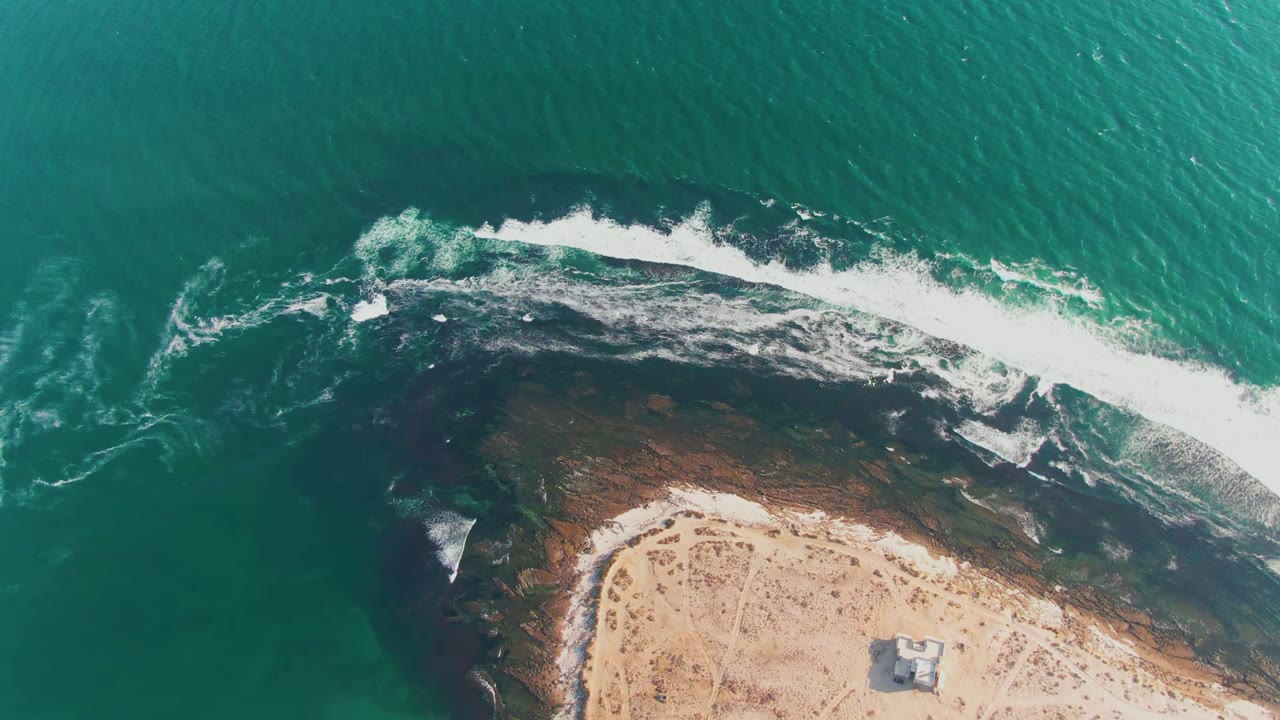 Top-down aerial frame at La Bocana showing turquoise water, a band of white surf over dark rock, sandy shore at the bottom, and no visible people.