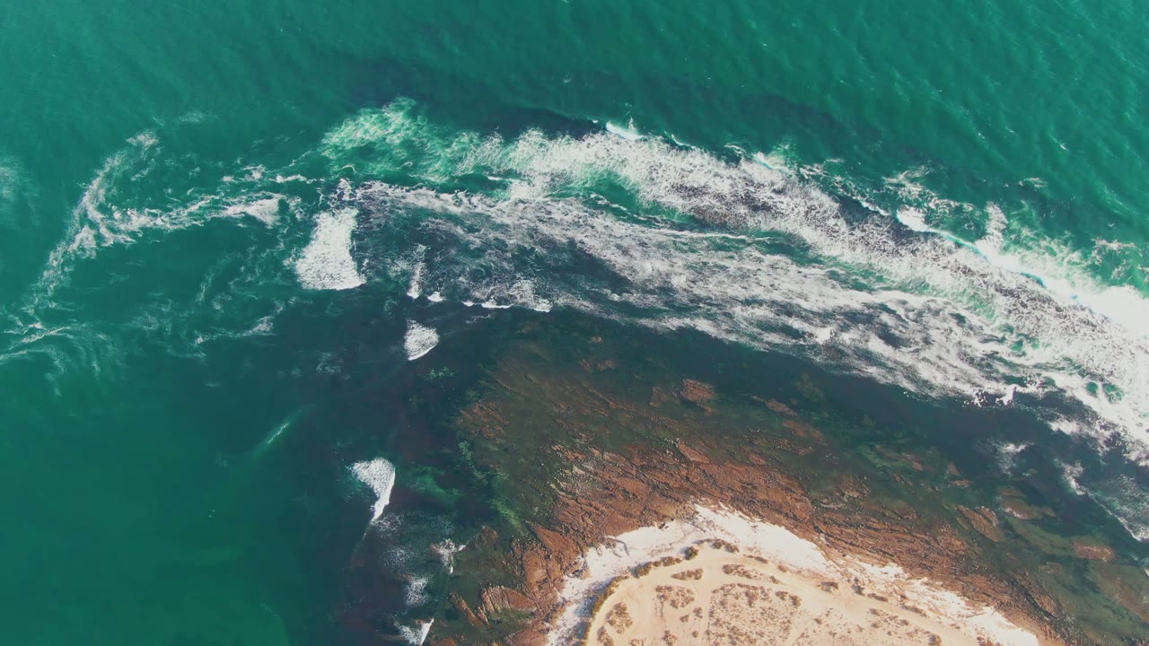 Aerial frame over La Bocana, Baja California Sur showing turquoise water, white foam, dark submerged rock, tan sand at the lower edge, and no visible people.