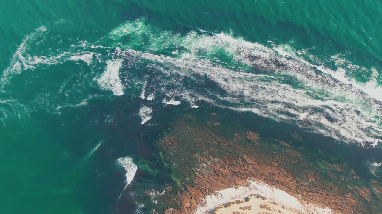 Aerial view at La Bocana showing a curling white foam line across green-blue water, exposed brown rock texture below, sand near the lower edge, and no visible people.