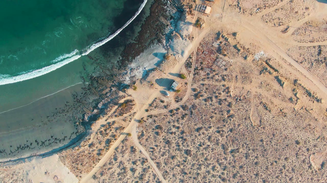 Overhead view of La Bocana, Baja California Sur, with green ocean at left, a white wave edge, rocky shoreline, pale sand tracks, dry scrubland, small structures, and no visible people.