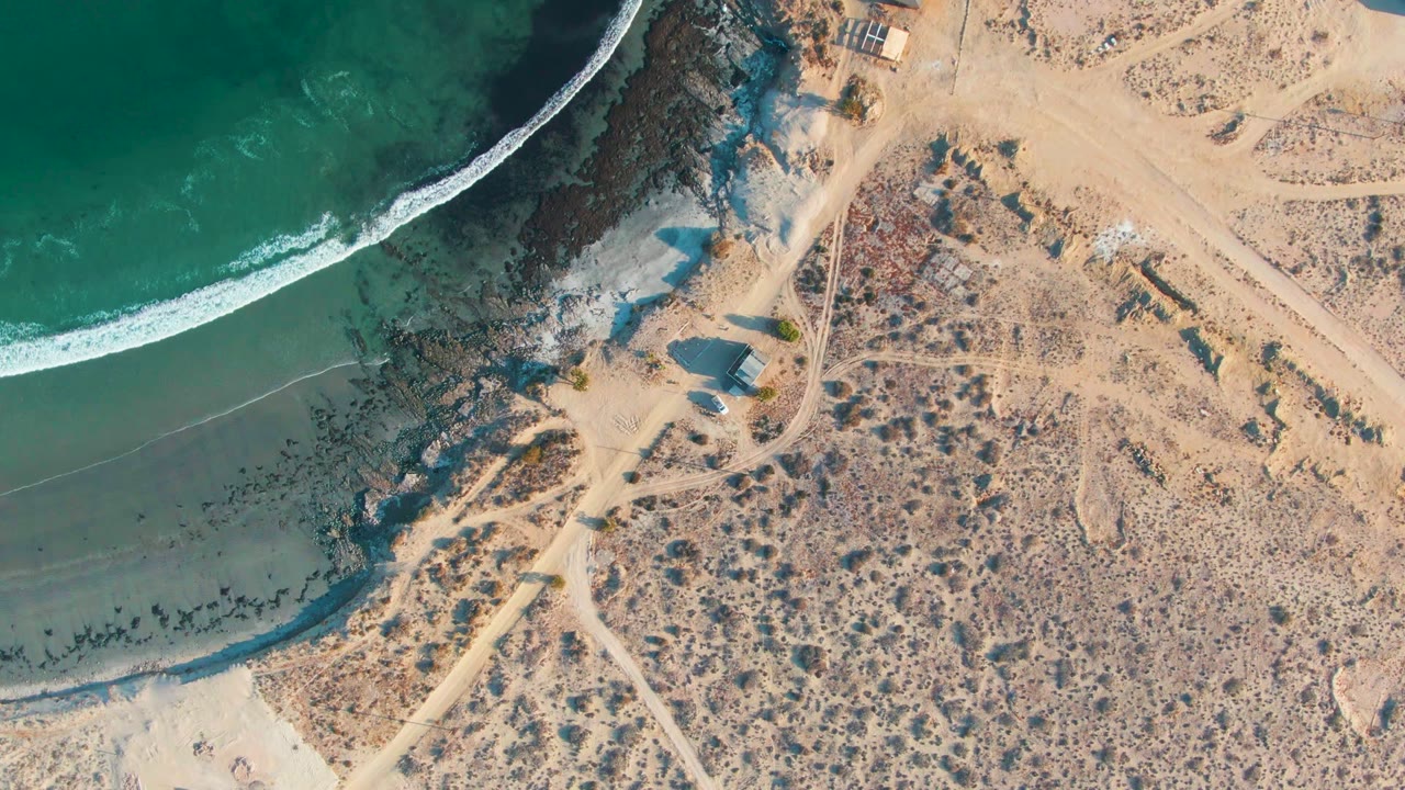 Overhead shoreline with surf line, rocky coast, sand tracks, and arid land.