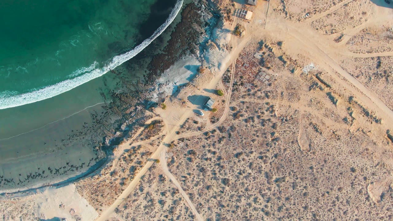 Top-down La Bocana coastline with green water filling the left side, a curved white break, dark shore rocks, tan dry land, intersecting dirt tracks, and no visible people.