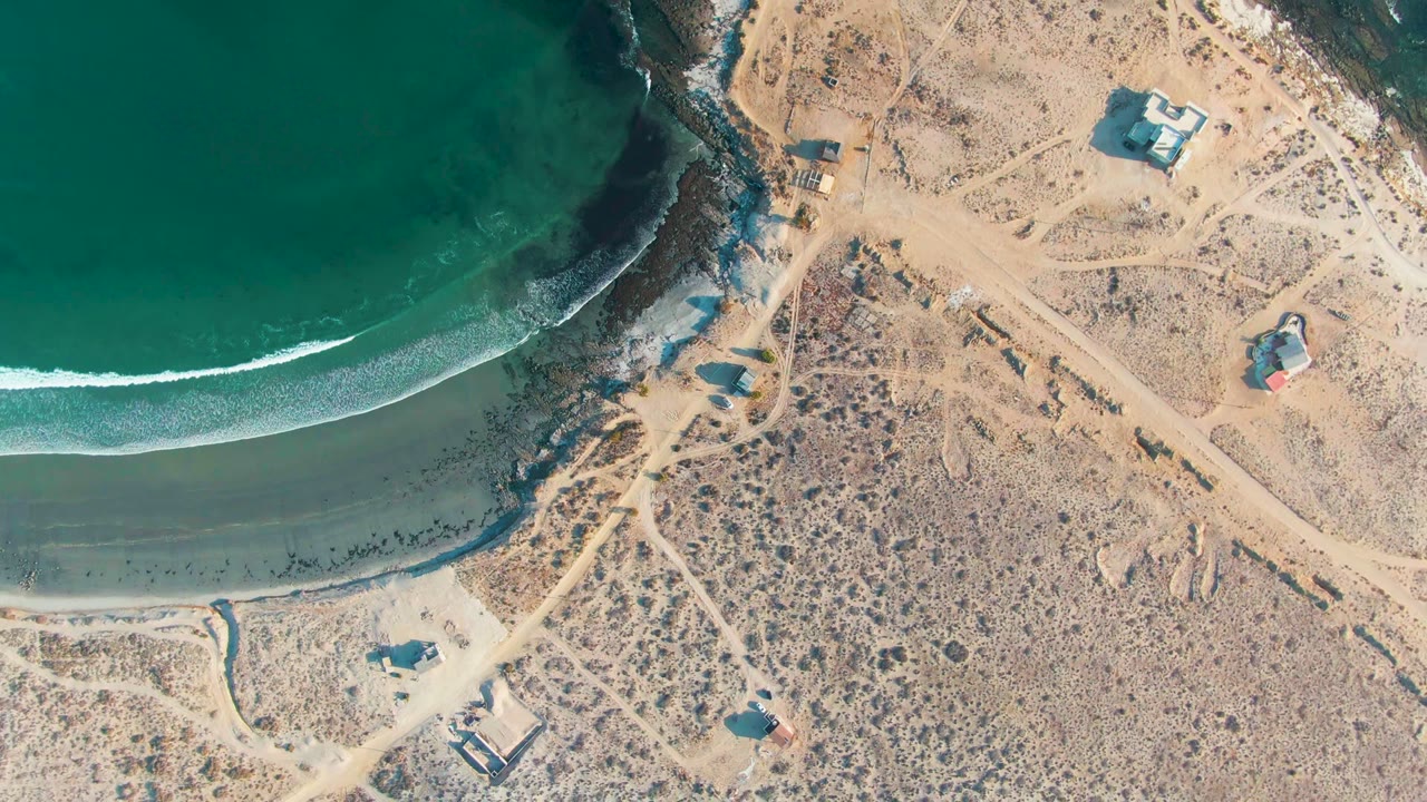 At 0:00, an overhead drone view in La Bocana, Baja California Sur shows turquoise water meeting a sandy beach beside dirt paths, scrubland, and small structures, with no visible people.