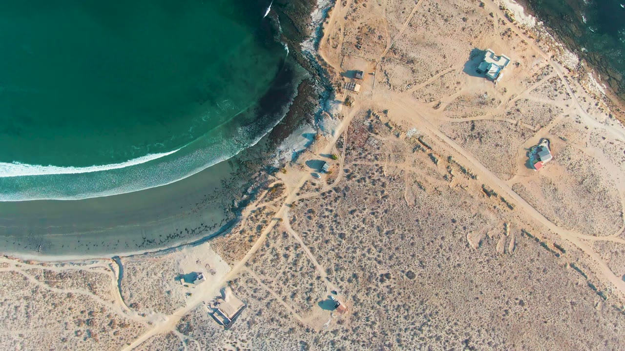 Aerial coastline with a curved wave line, dark wet sand, dirt tracks, and small buildings.