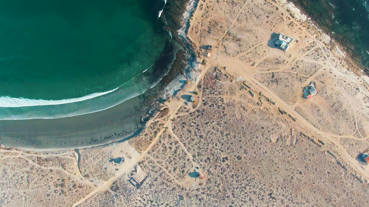 At 0:02, the overhead La Bocana frame shows white foam approaching the beach near a rocky point, with pale dirt paths and scattered small structures across the dry coastal land, and no visible people.