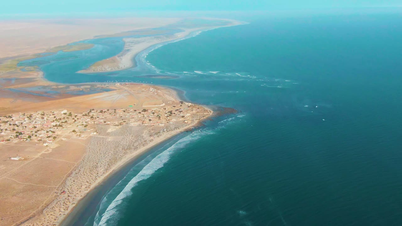 High aerial view over La Bocana, Baja California Sur, with a small sandy-shore settlement at lower left, a pale river mouth and sandbar beyond it, turquoise ocean to the right, and no visible people.