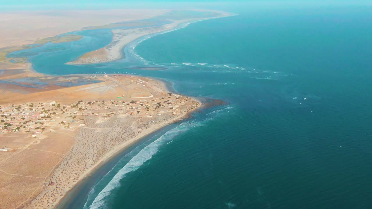 Aerial coastline with sandy settlement, river mouth, sandbar, and ocean surf.