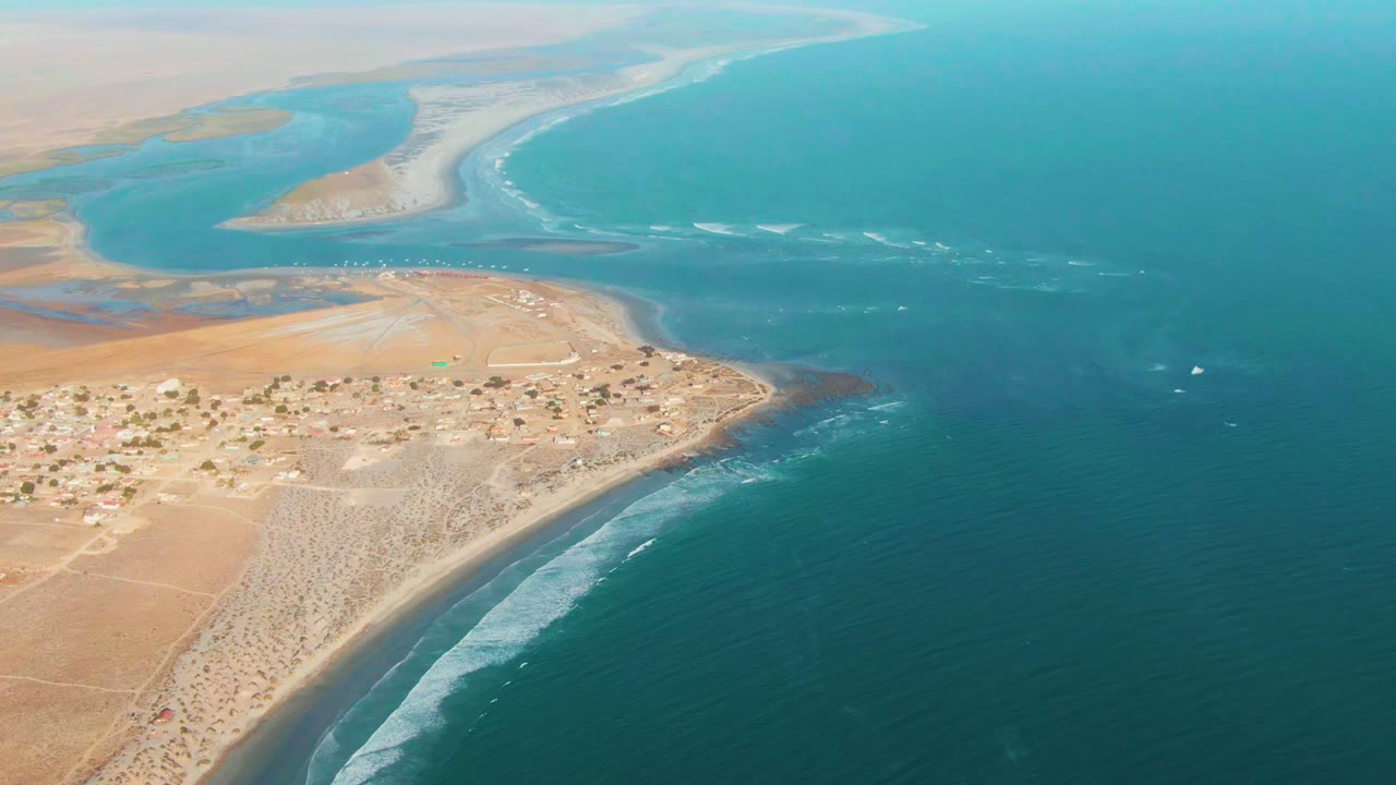 High oblique aerial view of La Bocana with tan arid land and huts near the lower left, white wave lines wrapping the sandy shoreline, deeper blue ocean on the right, and no visible people.