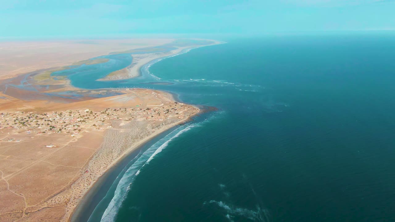 Aerial frame over La Bocana, Baja California Sur, showing tan desert land at left, a small coastal settlement, blue ocean at right, an estuary beyond the shore, and no visible people.