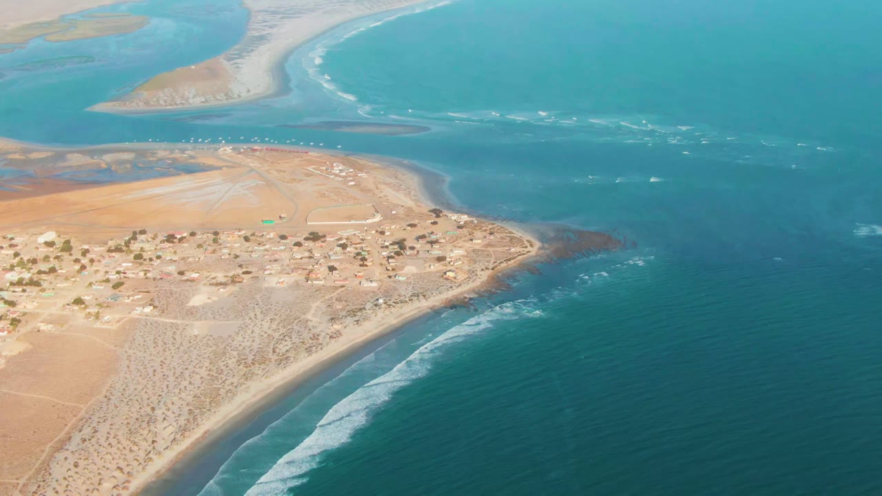 Aerial frame over La Bocana, Baja California Sur, showing a sandy settlement beside turquoise ocean, surf along the lower shore, pale sandbars beyond the inlet, and no visible people.