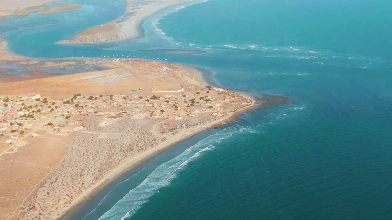 High aerial frame over La Bocana, Baja California Sur, with sandy village land at left, curved surf below, open blue water at right, and no visible people.