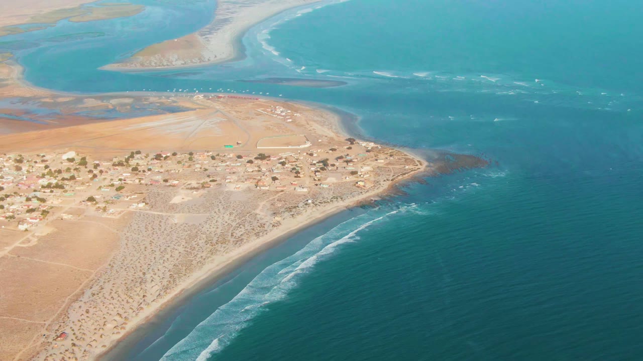 Wide aerial frame over La Bocana with the settlement centered left, white wave bands along the lower beach, turquoise shallows near the top, and no visible people.